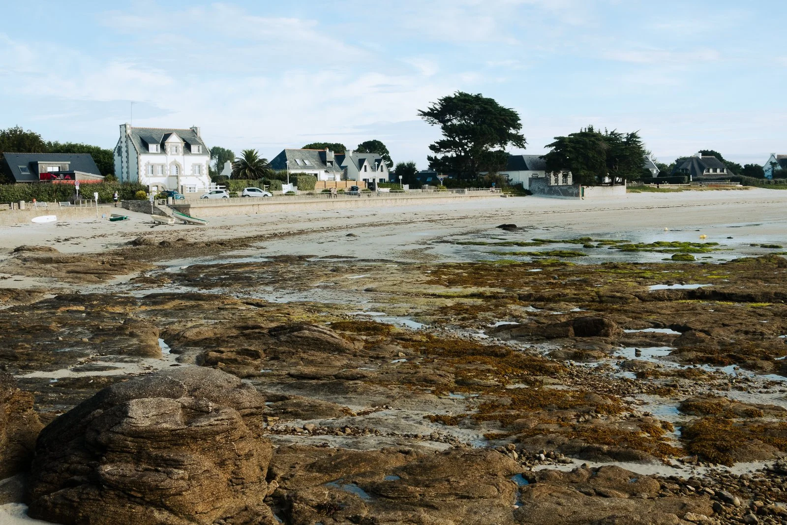 Plage avec rochers en premier plan, maisons et arbres au fond, ciel clair.