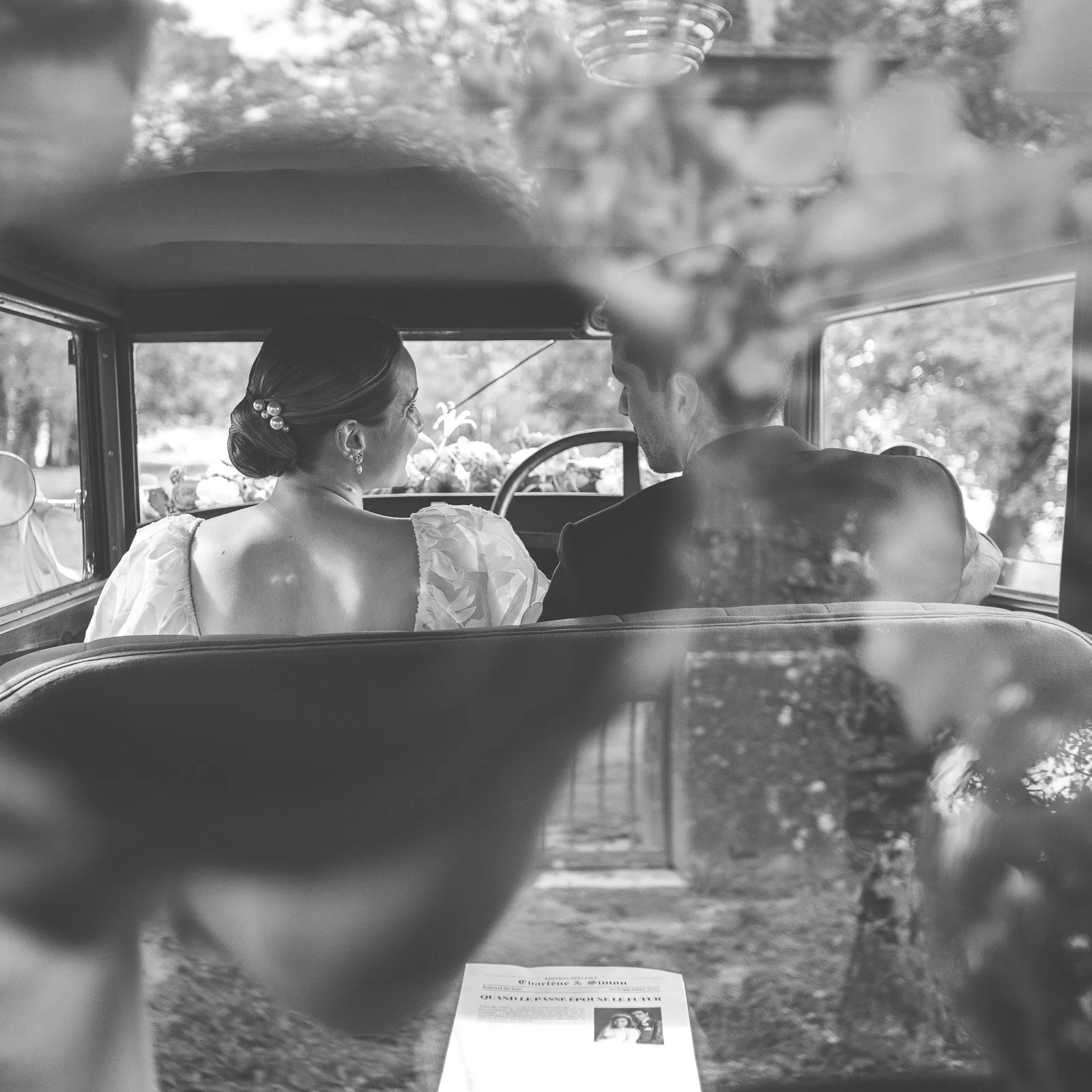 Un couple assis à l'arrière d'une voiture, une mariée et un marié, vue arrière, dans un stylisme noir et blanc.