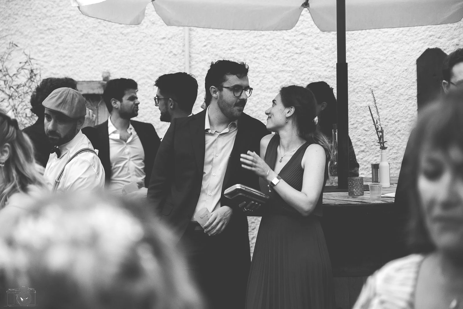Groupe de personnes conversant lors d'une fête ou d'un événement social en extérieur, sous un parasol, avec fond en mur clair et décor minimaliste.
