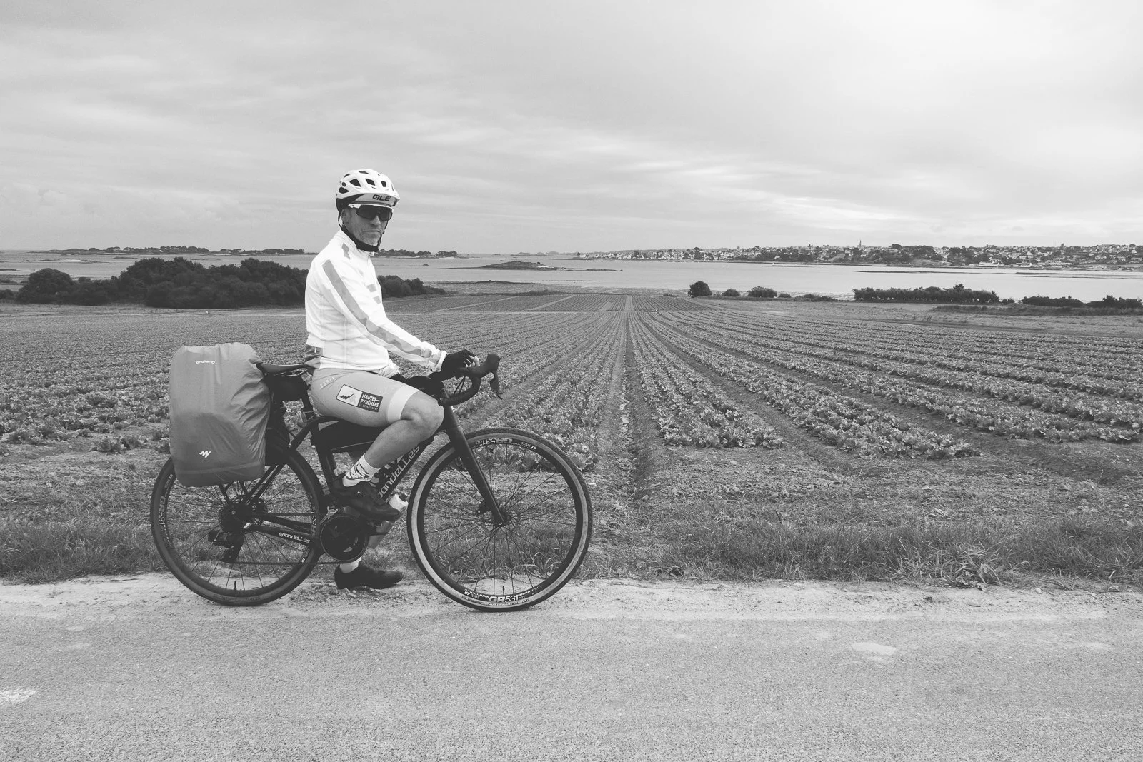 Homme en vélo avec un casque et des vêtements de cyclisme, en noir et blanc, sur un chemin de campagne avec un champ cultivé en arrière-plan.