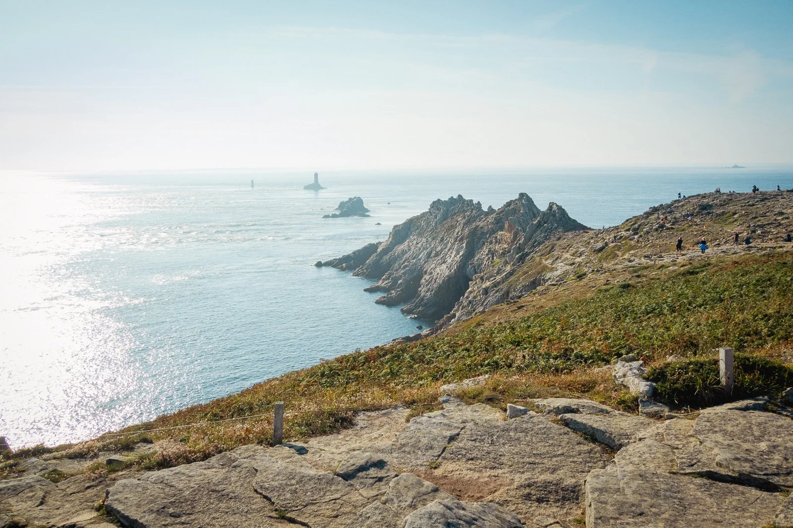 Paysage de falaises rocheuses au bord de la mer avec quelques personnes en promenade, sous un ciel clair.