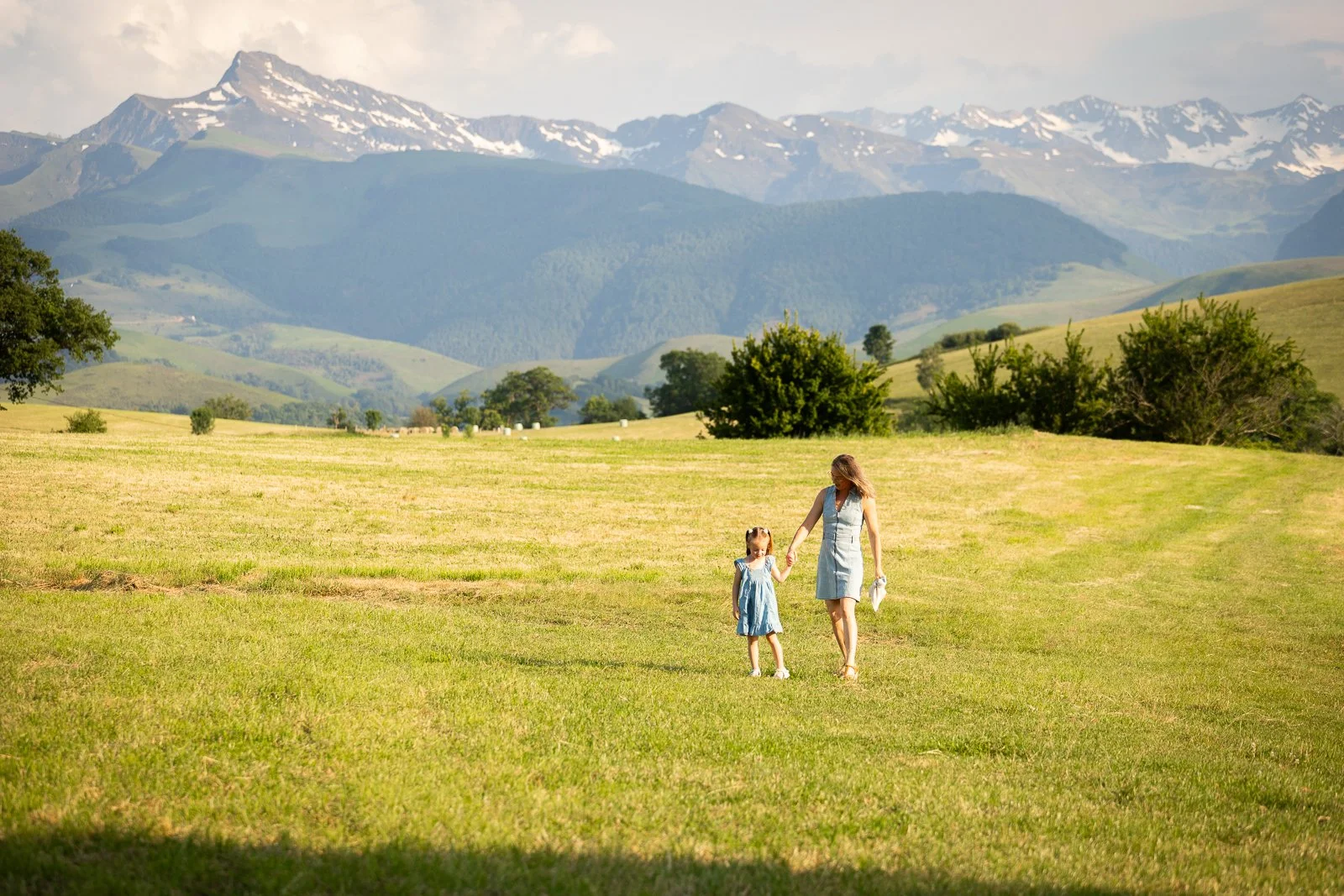 Une femme et une jeune fille marchent dans un champ vert avec des montagnes enneigées en arrière-plan, en journée ensoleillée.