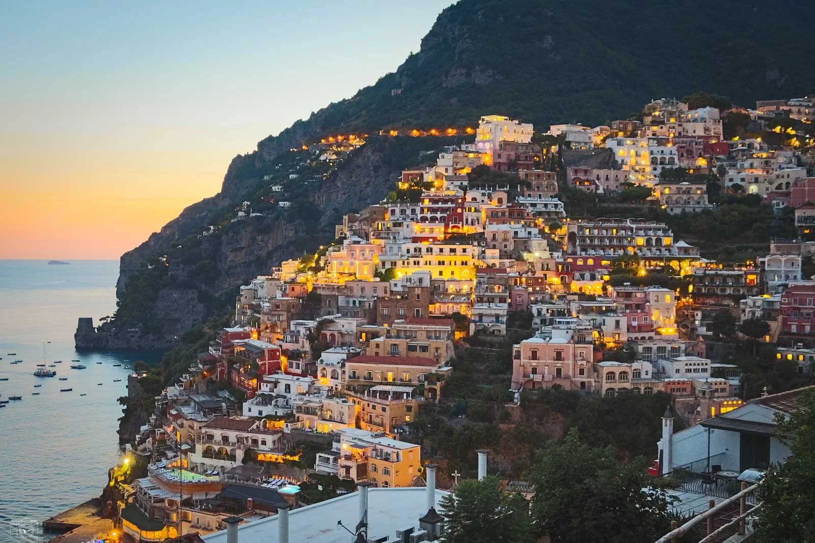 Vue à l'approche du village de Positano, en Italie, avec des maisons colorées suspendues sur les flancs d'une montagne, illuminées au crépuscule, surplombant la mer Méditerranée.
