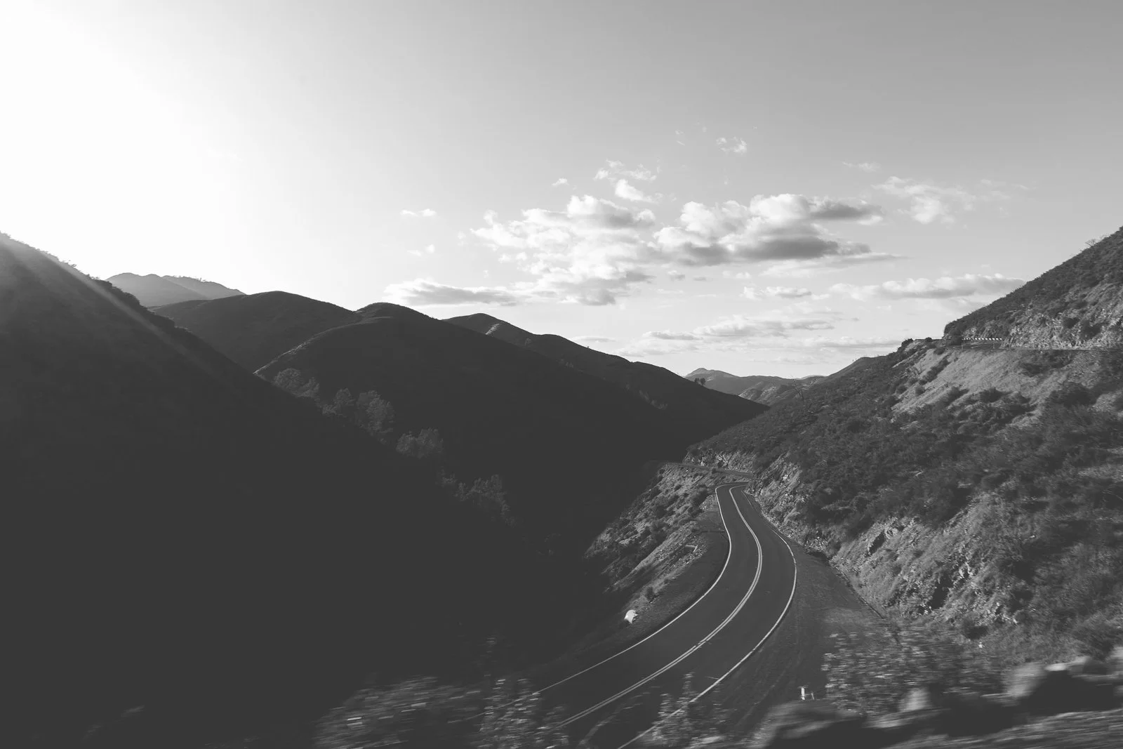 Une route sinueuse traverse un paysage montagneux, avec des collines escarpées recouvertes de végétation. Le ciel est partiellement nuageux.