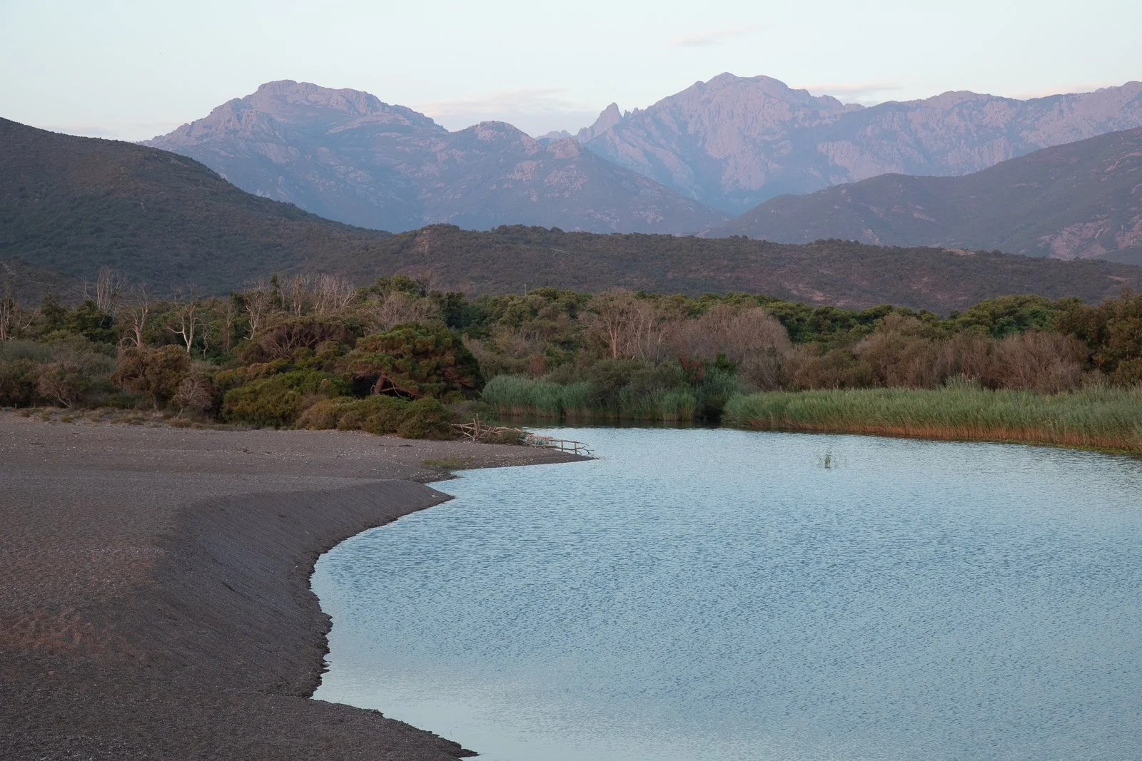 Un lac entouré d'arbustes et d'arbres, avec des montagnes en arrière-plan au coucher du soleil.