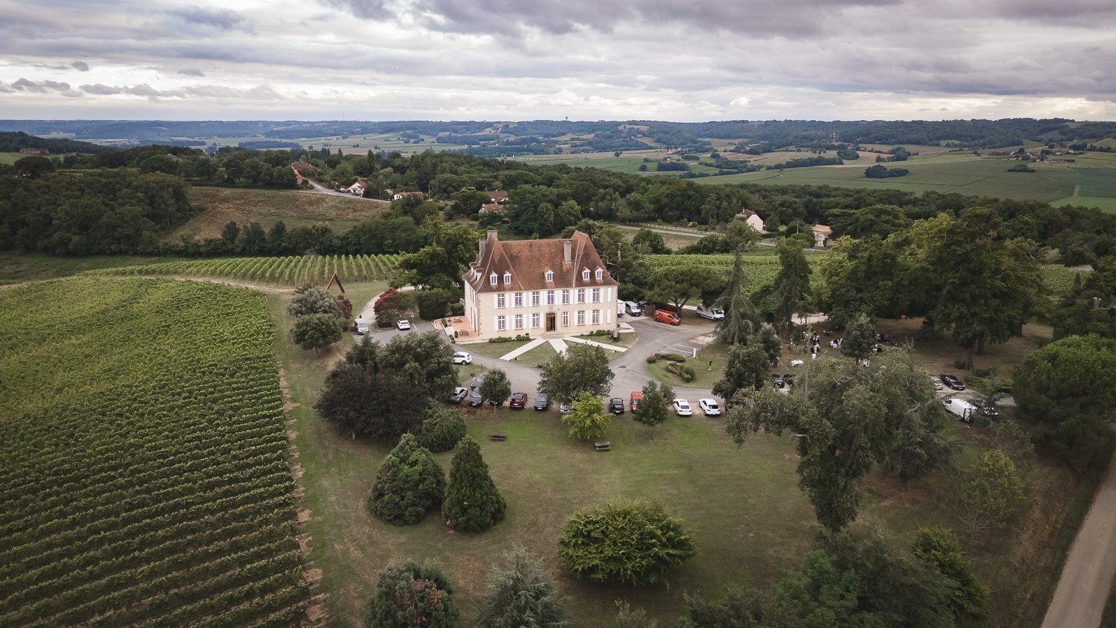 Vue aérienne d'un grand manoir blanc entouré de jardins, de nombreux arbres et de champs vallonnés dans une région rurale, sous un ciel nuageux.
