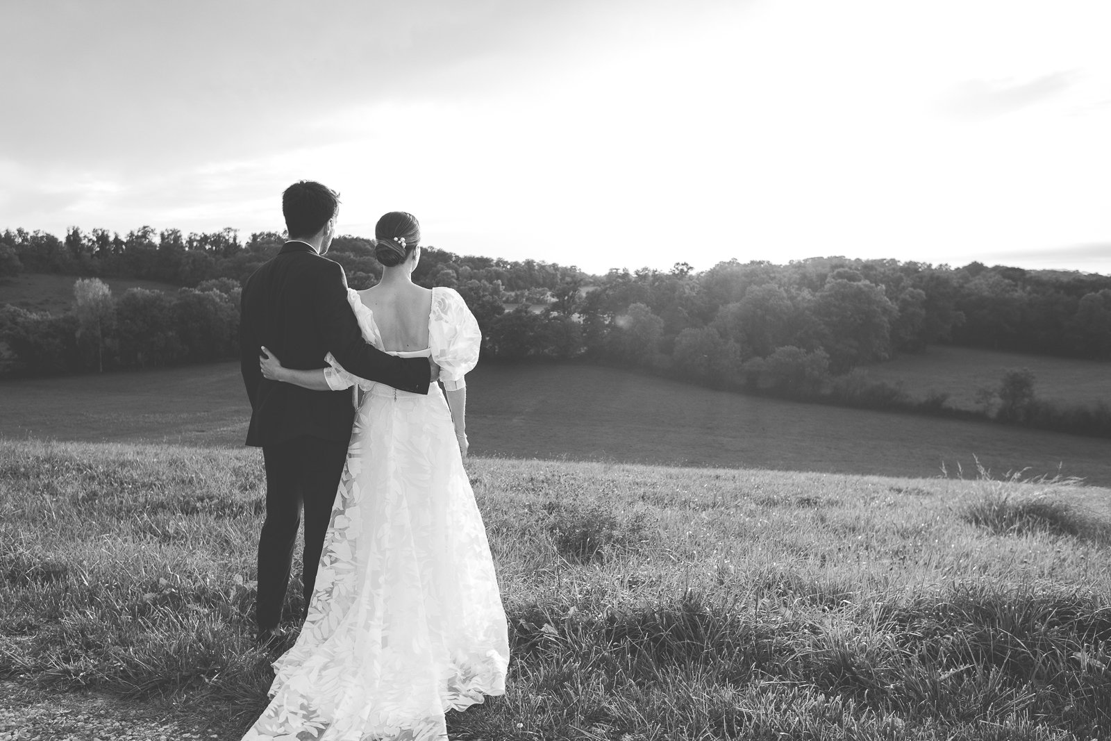 Un couple de mariés regardant un paysage bucolique avec des champs et des arbres, photographiés en noir et blanc.