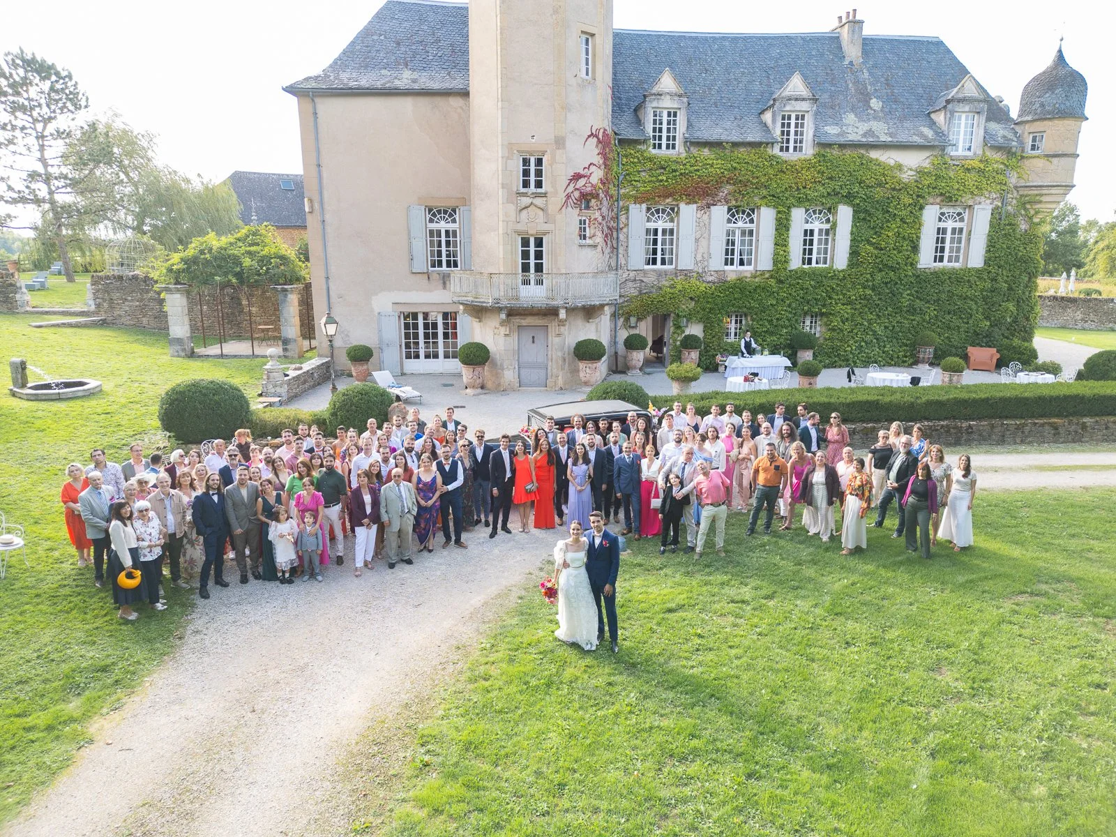Un groupe nombreux de personnes posant pour une photo devant un vieux manoir lors d'un mariage. La mariée en robe blanche et le marié en costume noir se tiennent à l'avant avec un bouquet de fleurs.
