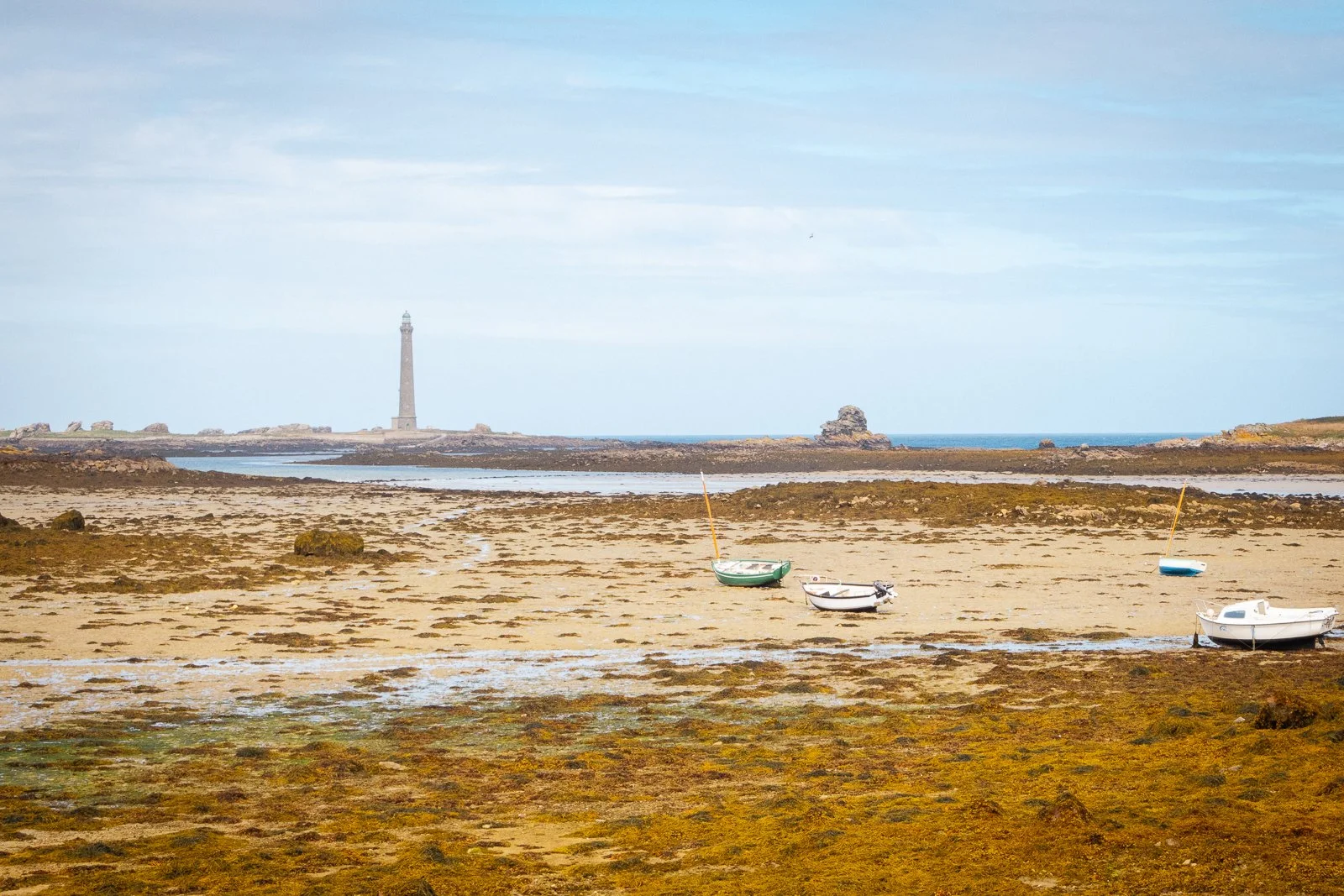 Vue de plages avec des bateaux à marée basse, un phare en arrière-plan, ciel partiellement nuageux.