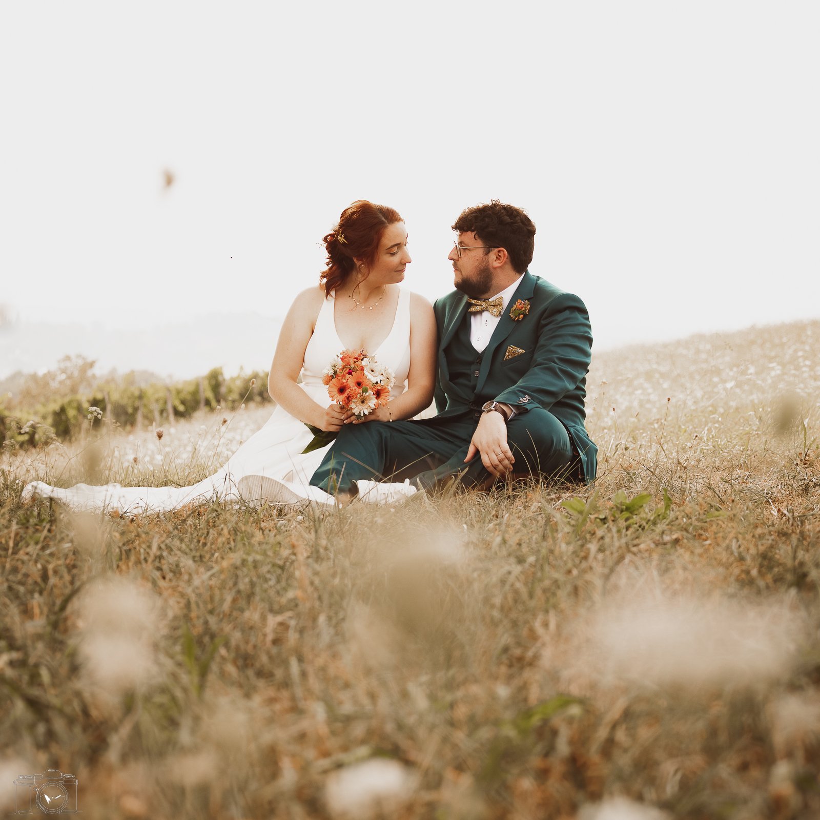 Un couple de mariés assis dans un champs lors d'un mariage, la femme tient un bouquet de fleurs et regarde son mari, tous deux en tenue de mariage.