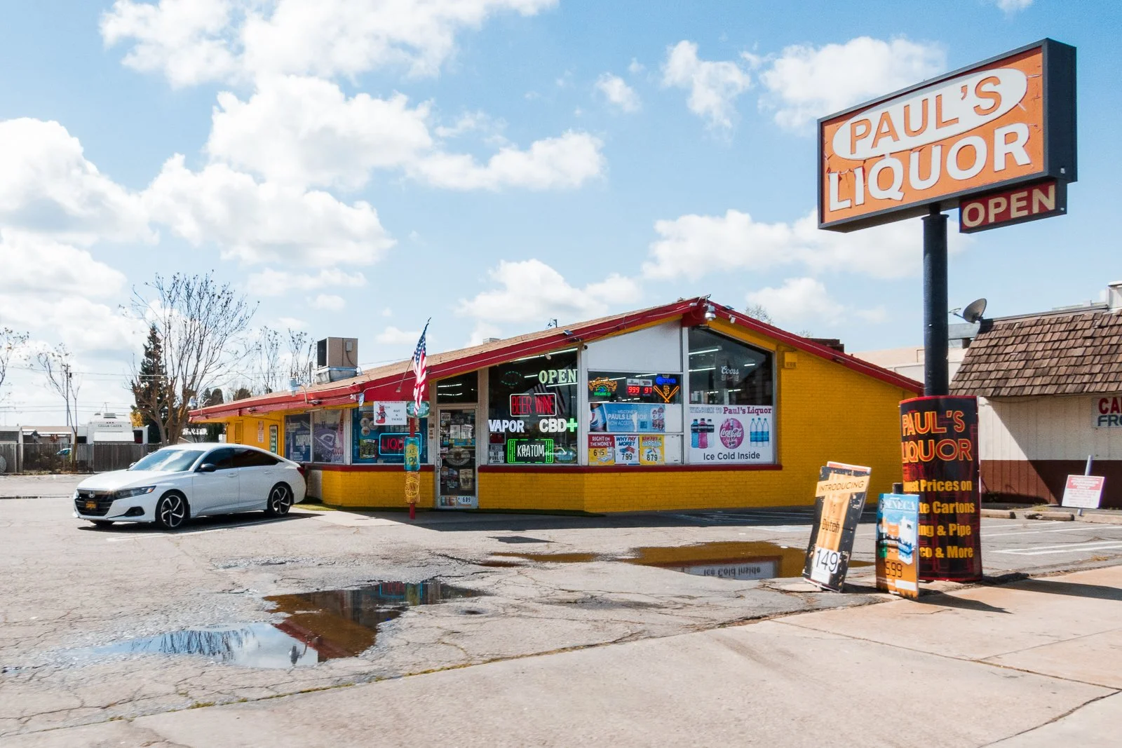 Magasin appelé Paul’s Liquor avec enseignes lumineuses, voiture blanche garée devant, panneau indiquant des promotions et produits divers, ciel partiellement nuageux.