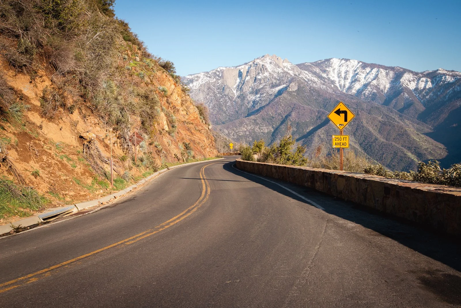 Une route sinueuse en montagne avec un panneau de signalisation indiquant un virage à gauche dans environ 250 pieds, avec des montagnes enneigées en arrière-plan.