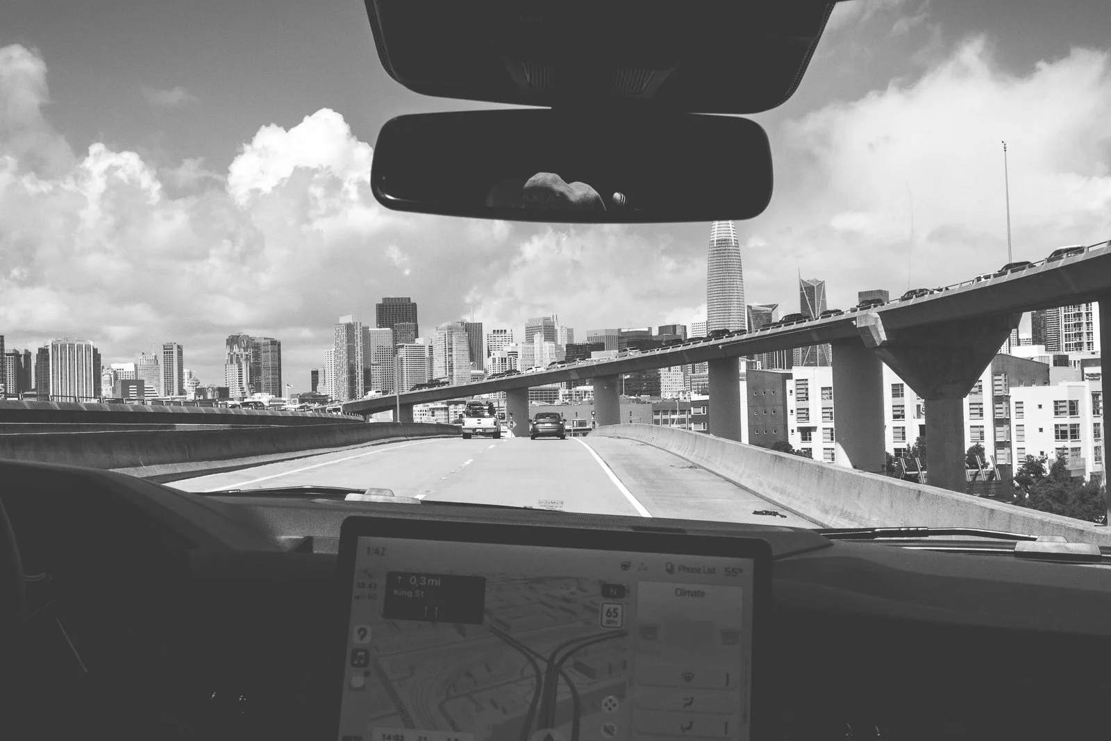 La vue depuis l'intérieur d'une voiture en conduite sur une autoroute de San Francisco, avec le skyline de la ville visible en arrière-plan, en noir et blanc.