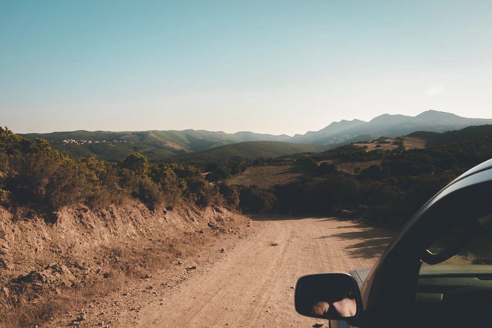 Vue d'une route de terre entourée de haies et de collines avec montagnes à l'horizon, côté droit d'un véhicule en conduisant.