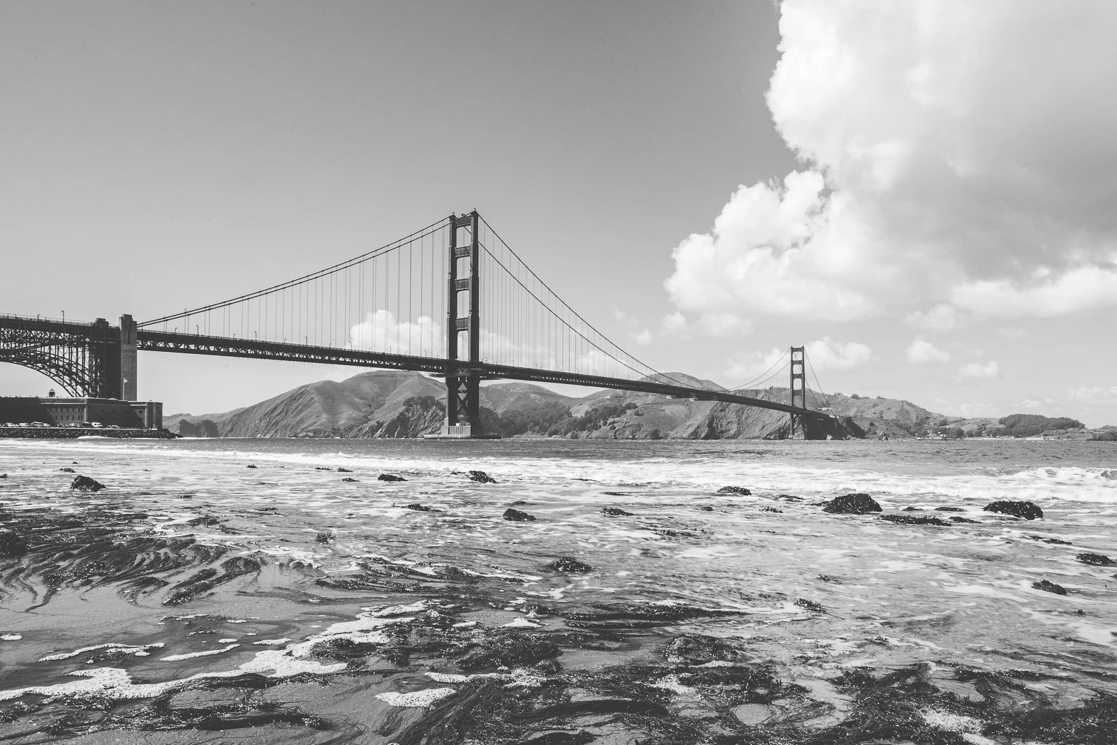 Le pont Golden Gate vu de la plage, avec des montagnes en arrière-plan et des nuages dans le ciel, en noir et blanc.