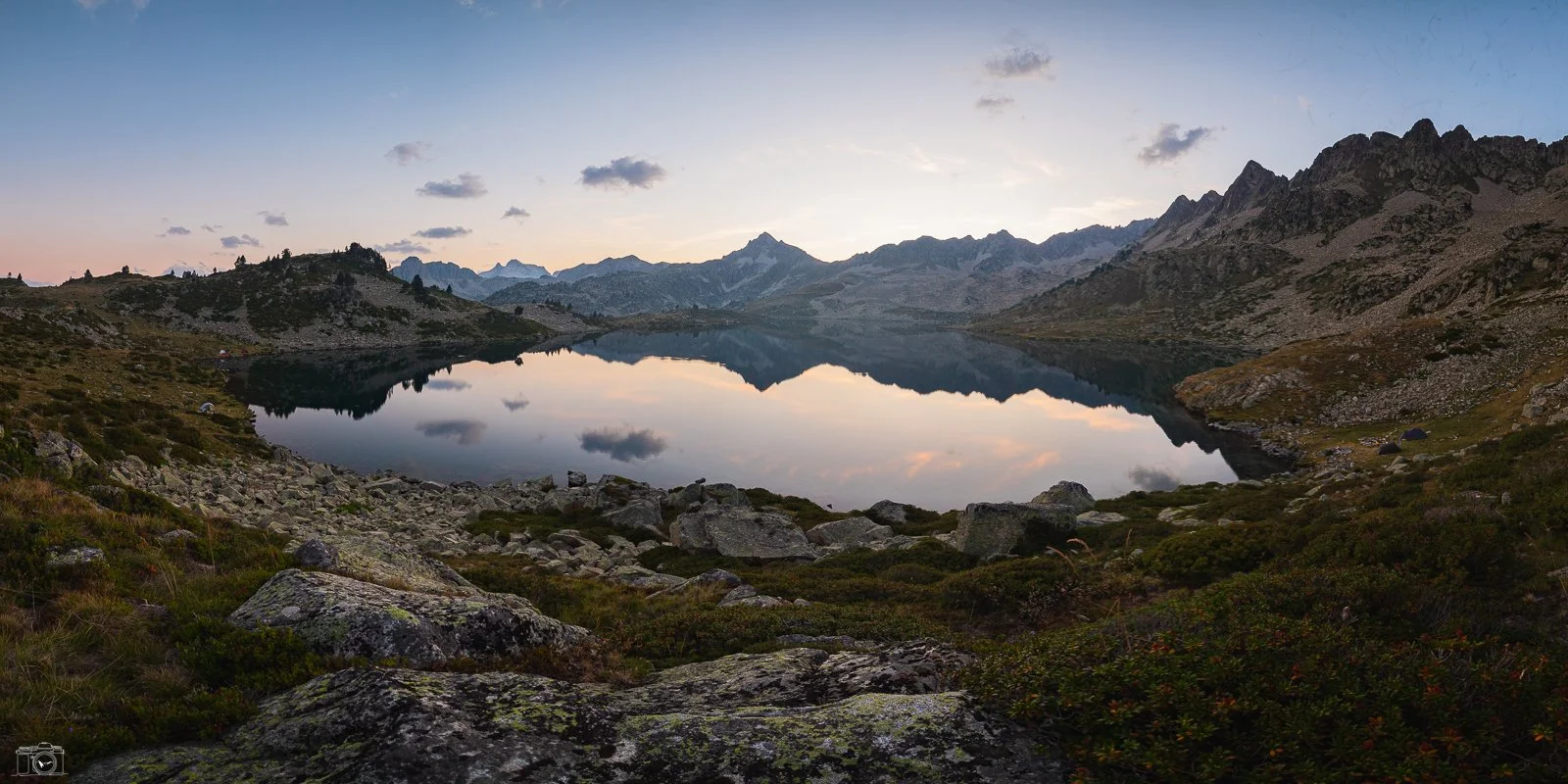 Un lac de montagne calme reflétant les pics enneigés environnants, avec un ciel au coucher du soleil et des rochers et végétation au premier plan.