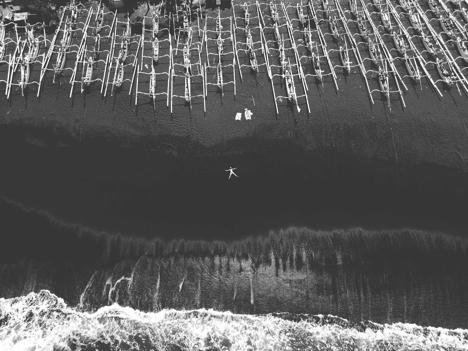 Photo en noir et blanc d'une plage avec des structures de surf ou de paddle alignées, vue du ciel. Au centre, un cercle formé par une personne allongée sur la plage.