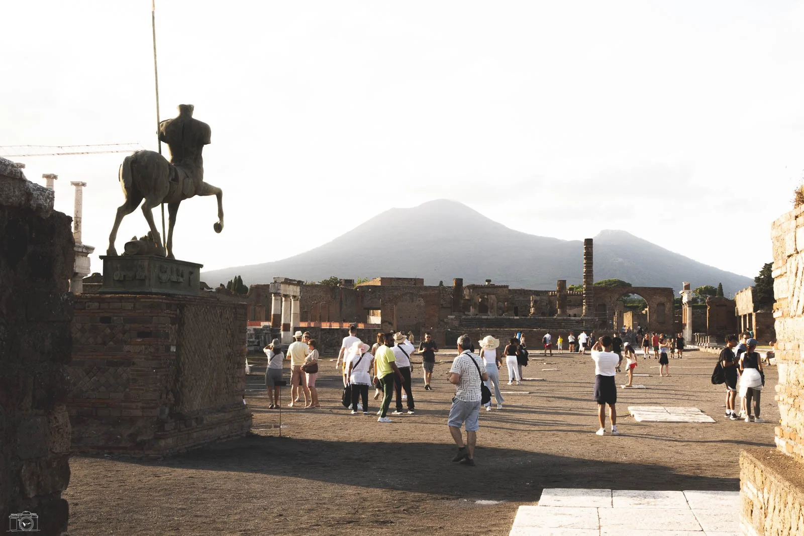 Ruines antiques avec de nombreux touristes, une statue équestre en premier plan, un volcan en arrière-plan, et un arc antique