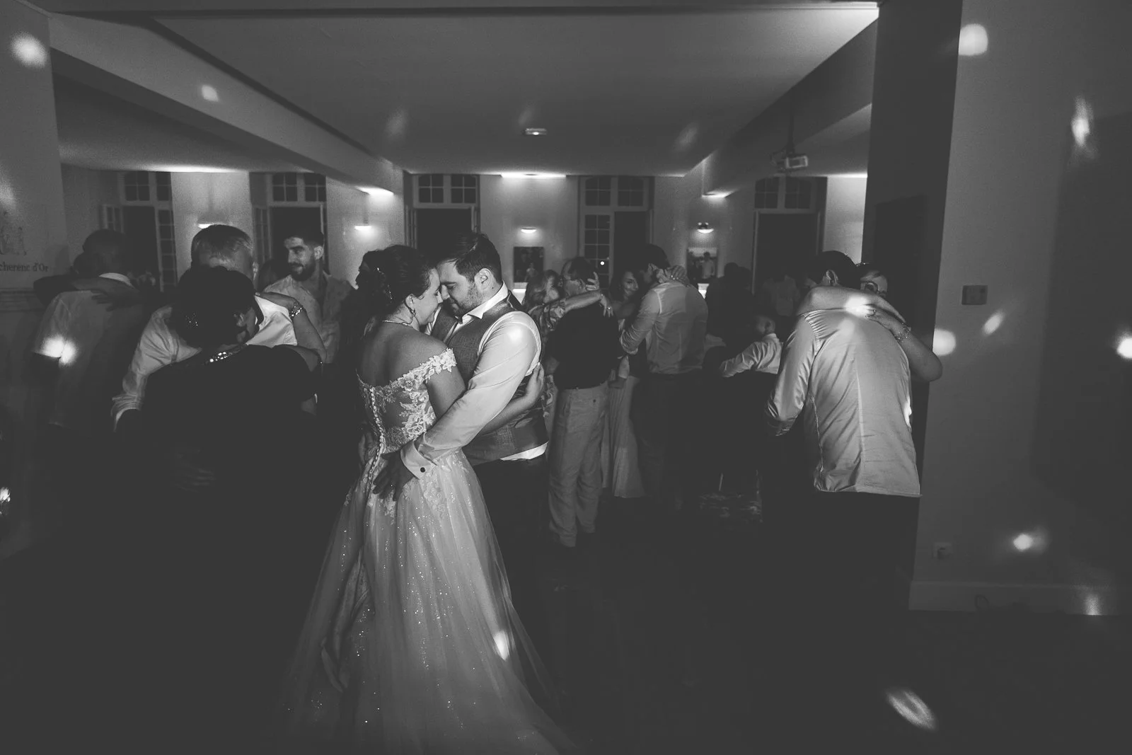 Une scène de mariage avec plusieurs couples dansant dans une salle. En premier plan, un couple de mariés, la mariée en robe de princesse et le marié en costume, dans une posture intime de danse.