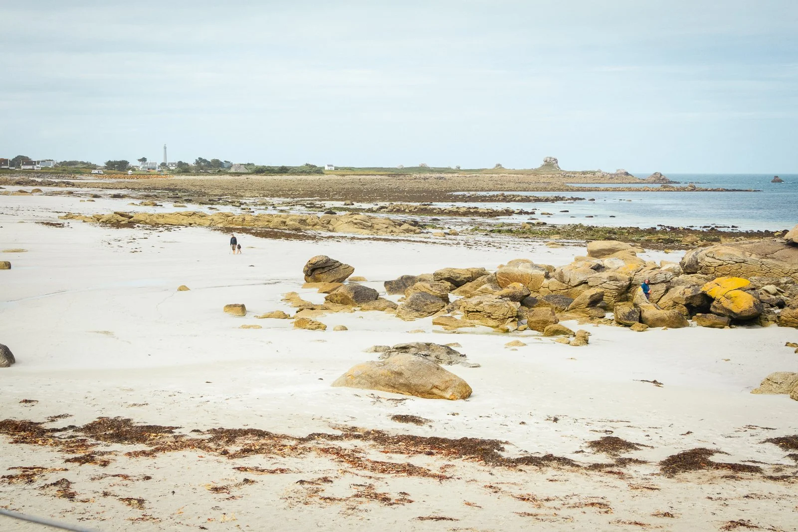 Plage avec sable blanc, rochers, deux personnes marchant et une autre assise, au loin un phare et des maisons près de la côte.