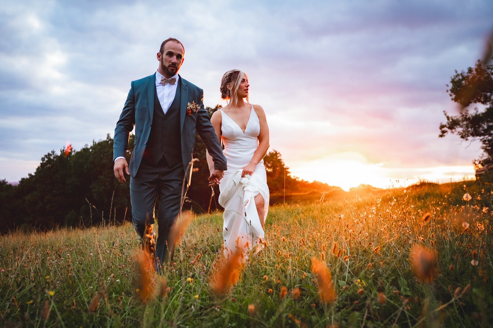 Un couple de mariés marche dans un champ en fleurs lors d'un coucher de soleil, la femme en robe blanche et le homme en costume bleu avec un nœud papillon.
