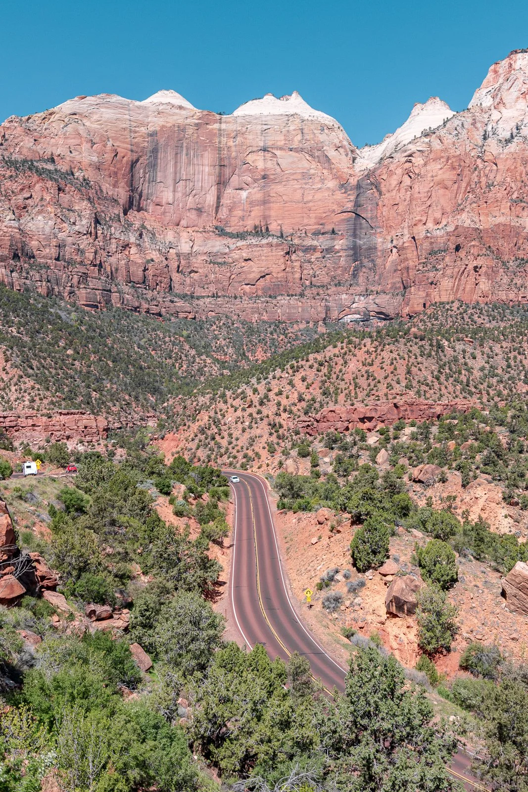 Une route sinueuse traverse un paysage montagneux avec des roches rouges et des arbres verts, sous un ciel bleu clair.
