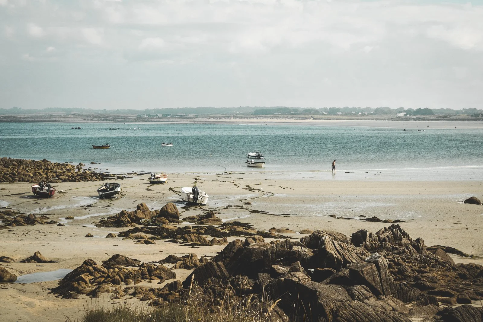 Plage avec des bateaux échoués sur le sable, des rochers en premier plan et la mer calme sous un ciel nuageux.