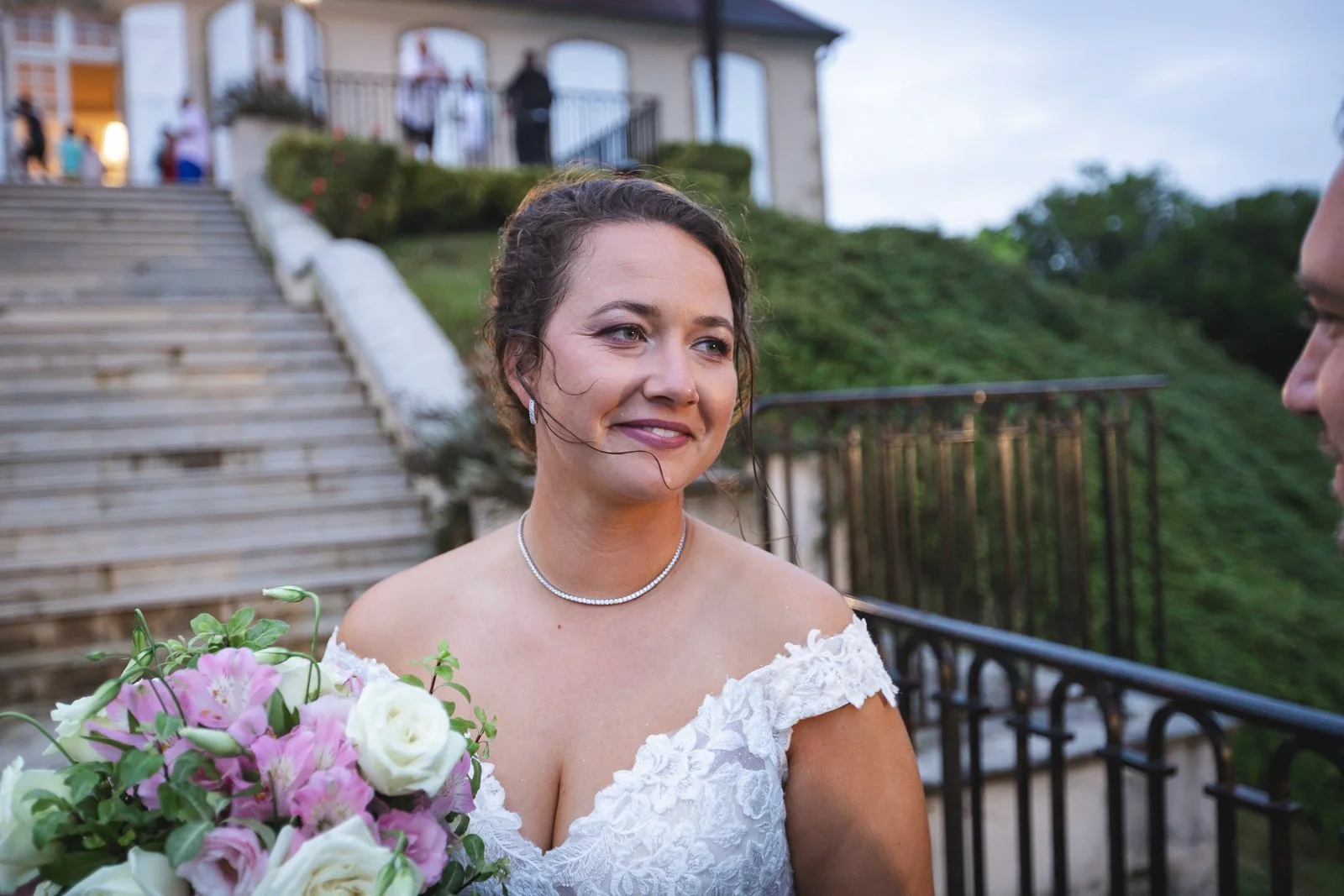 Une femme en robe de mariage tient un bouquet de fleurs roses et blanches lors d'une cérémonie en extérieur devant un bâtiment avec des escaliers, un garde-corps en fer et des invités en arrière-plan.