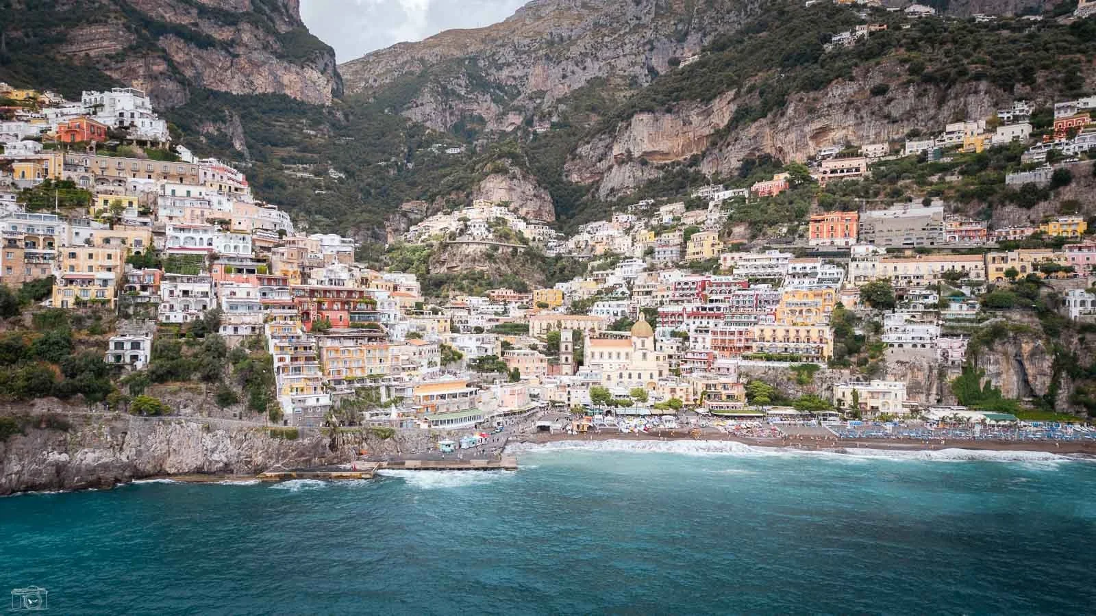 Vue de la côte italienne, montrant des maisons colorées en pente sur une falaise surplombant la mer turquoise, avec des montagnes en arrière-plan.