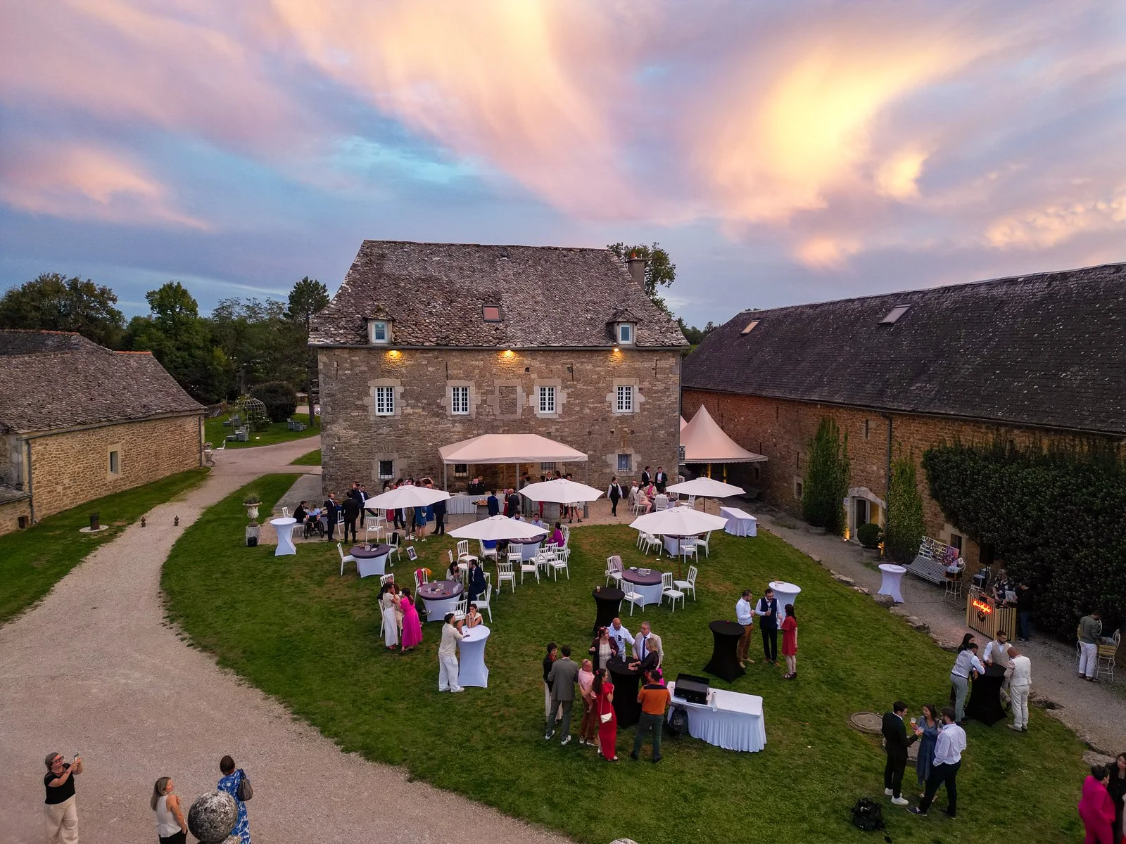 Une réception ou un mariage en plein air dans un vieux bâtiment en pierre, avec des invités, des tables avec chapeaux de soleil, et un ciel coloré au coucher du soleil.