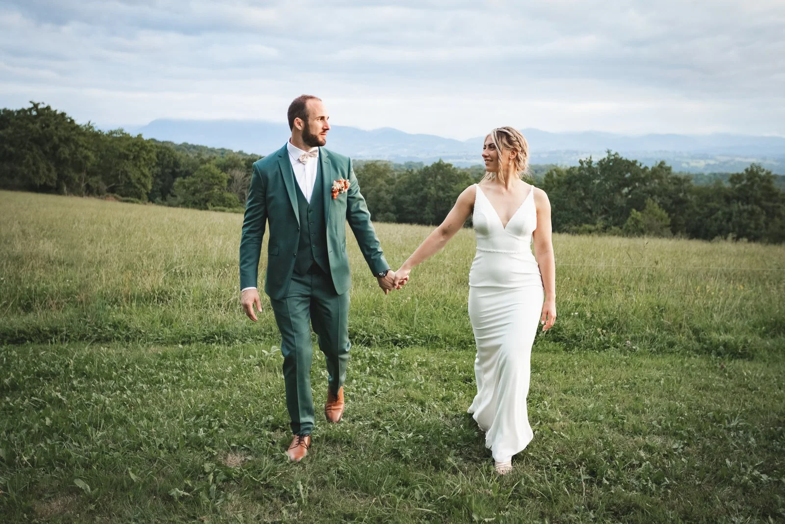 Un couple en mariage marchant dans un champ vert avec un paysage de montagnes en arrière-plan, la femme porte une robe blanche et l'homme un costume vert avec une veste et un pantalon ajustés.