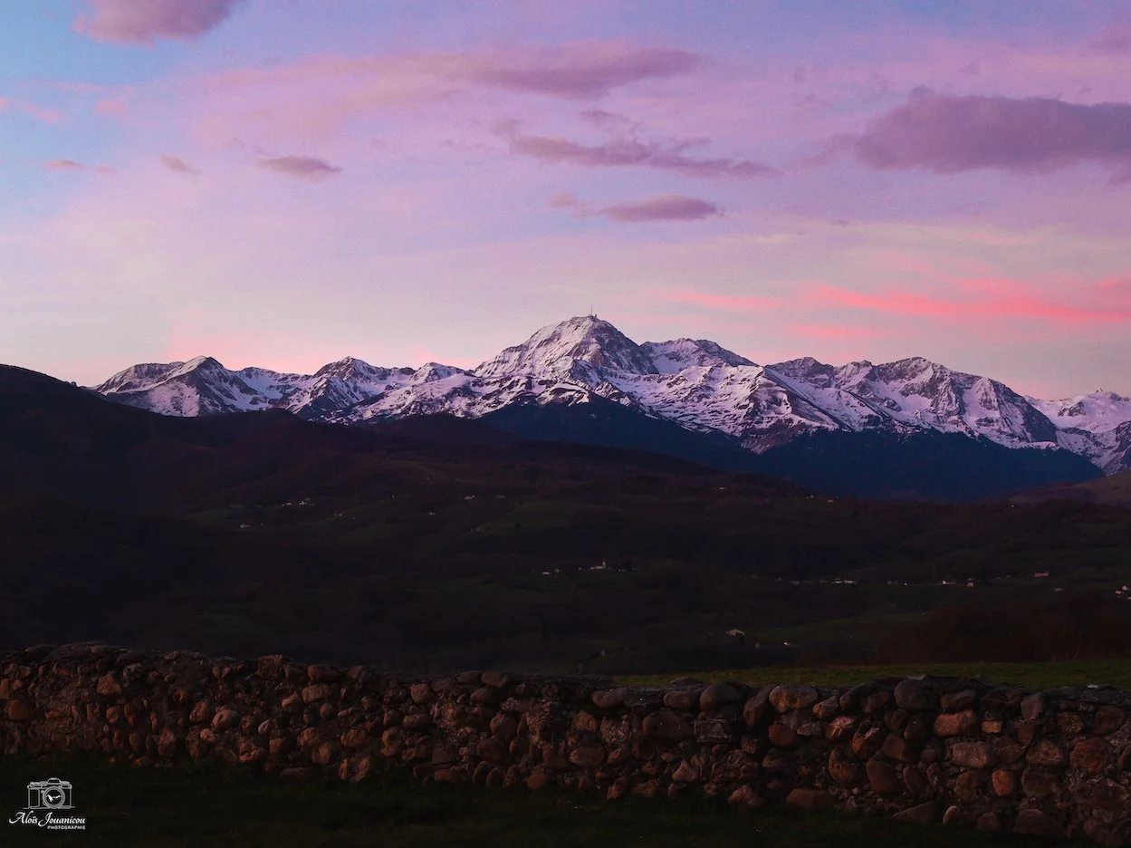 Montagnes enneigées au coucher du soleil avec un ciel rose et violet, un mur de pierres au premier plan, paysage paisible