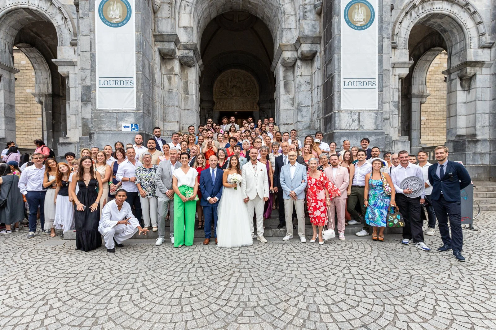 Groupe de personnes posant pour une photo de mariage devant une grande porte d'édifice en pierre à Lourdes, avec des panneaux indiquant Lourdes, plusieurs mariés en tenue blanche, différentes tenues colorées, et des invités souriants.