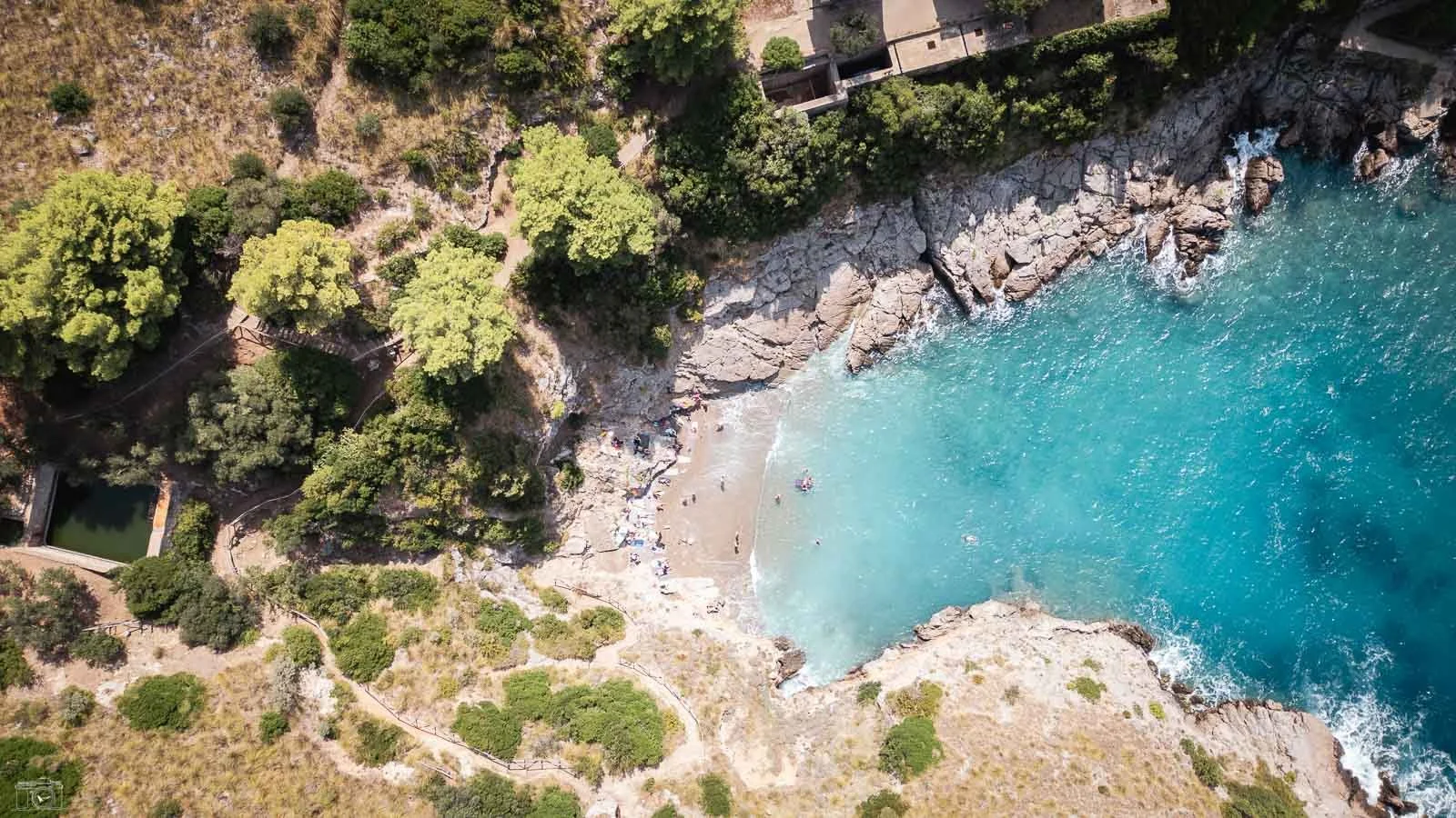 Vue aérienne d'une petite plage entourée de rochers et d'eau turquoise, avec des arbres verts le long du rivage.