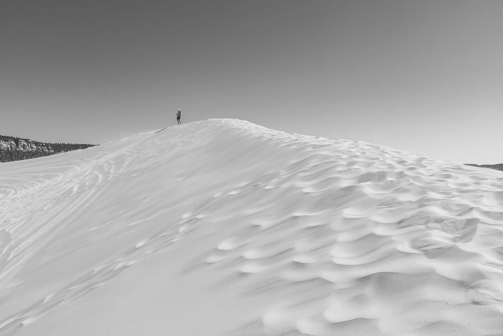 Une silhouette humaine se tient au sommet d'une dune de sable ou de neige, sous un ciel clair.