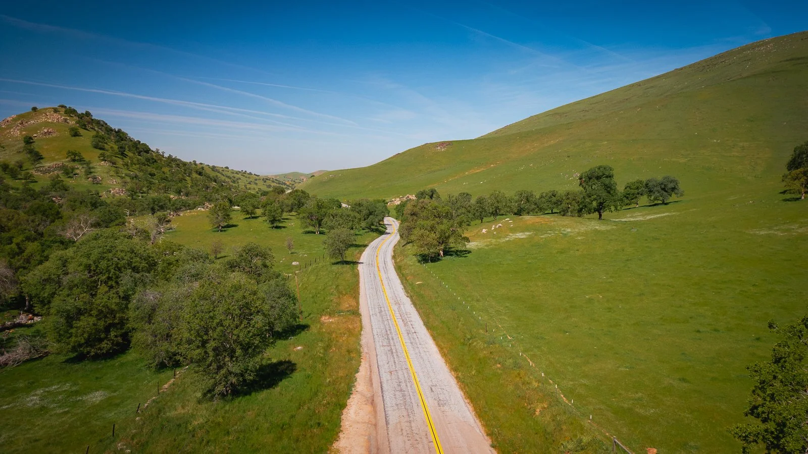 Route de campagne sinueuse au milieu de collines verdoyantes et de champs ouverts, sous un ciel bleu clair avec quelques traînées de nuages.