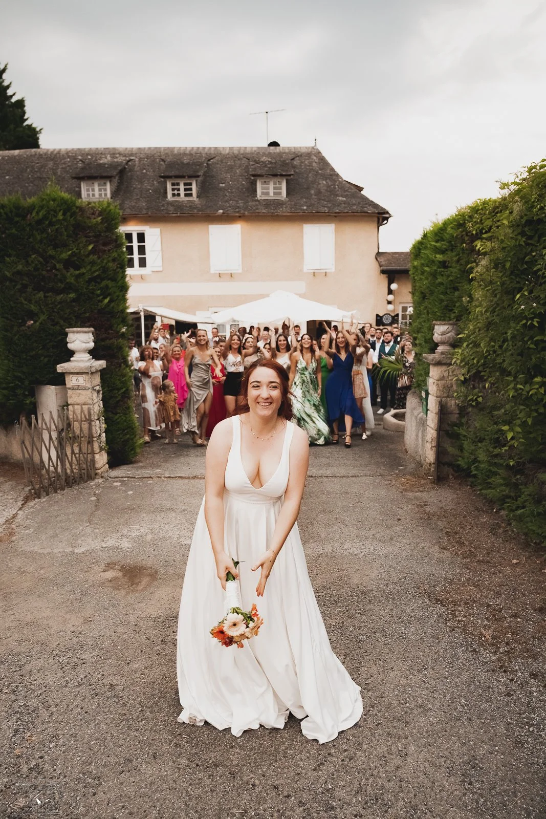 Une mariée souriante en robe blanche, tenant un bouquet, devant une foule de personnes en tenue élégante lors d'une célébration en plein air.