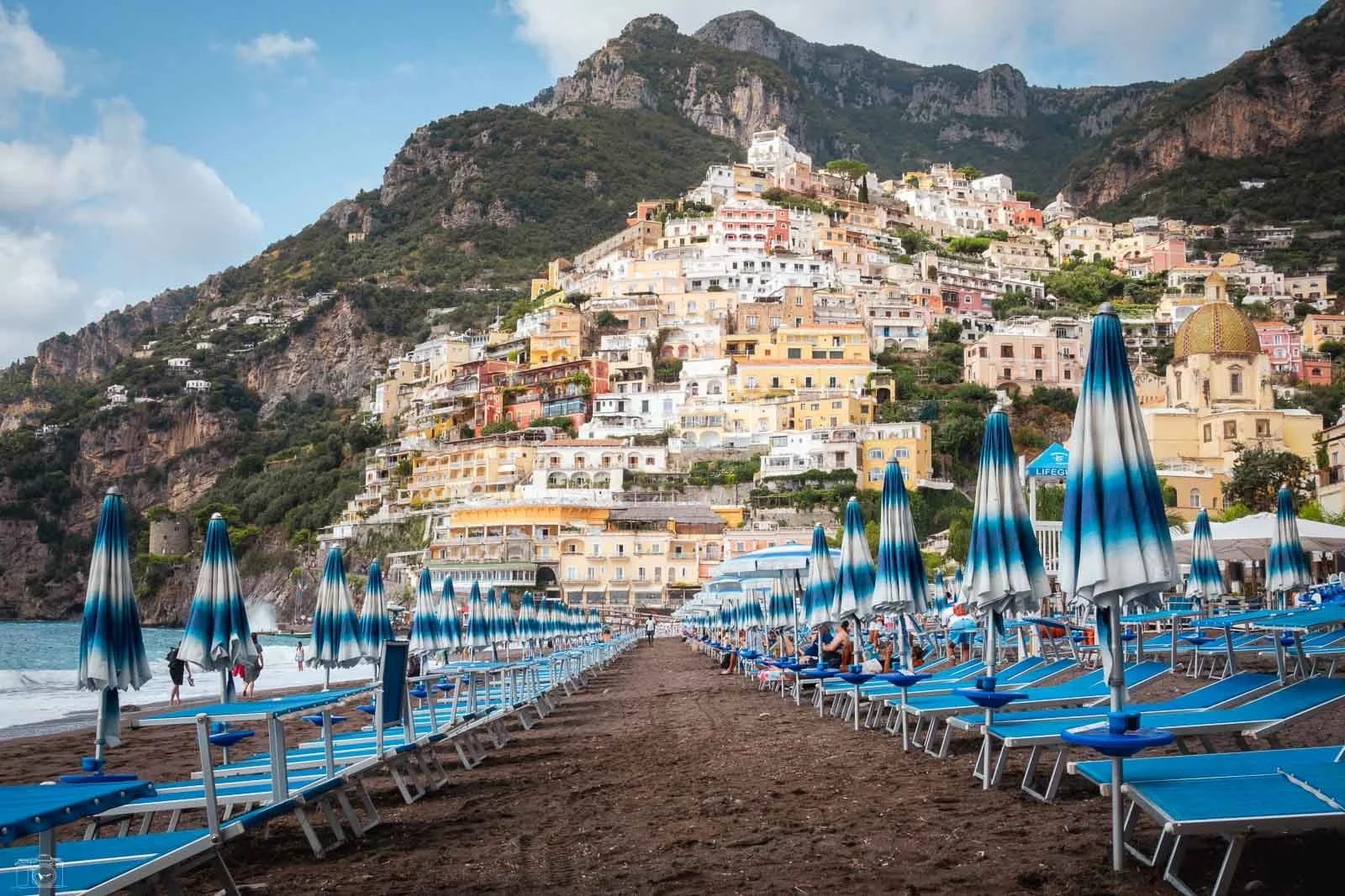 Plage avec des parasols bleus et des chaises longues face à une ville colorée construite sur une colline avec des maisons peintes de couleurs pastel