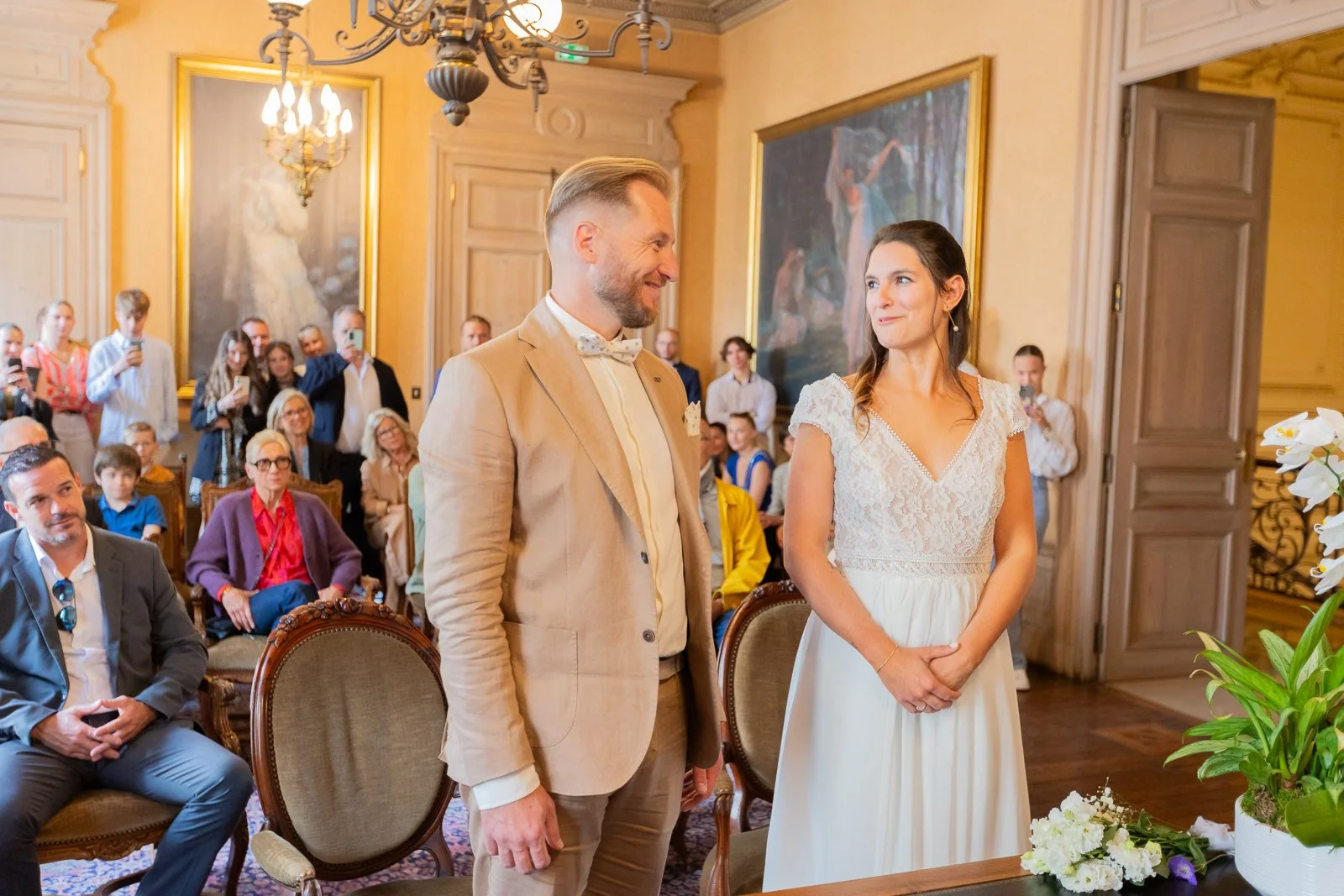 Un couple lors d'un mariage, la mariée porte une robe blanche à manches courtes et le marié un costume beige avec un nœud papillon blanc. Ils se regardent tendrement dans une salle élégante avec des invités en toile de fond, certains prennent des pho