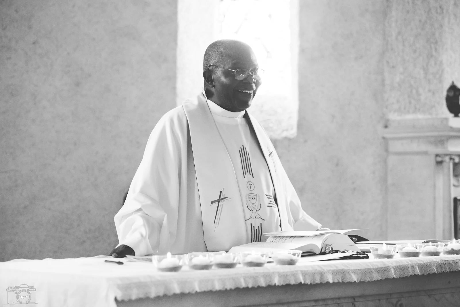 Un prêtre souriant, habillé d'une robe blanche avec des symboles religieux, se tenant à une table avec un livre ouvert et des petites bougies.