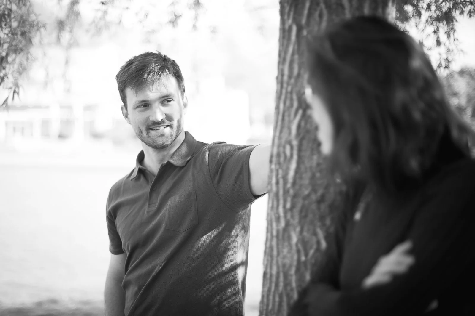 Deux personnes discutent à l'extérieur, avec un arbre entre elles. La personne à gauche est un homme avec une barbe et un t-shirt à col polo, souriant. La personne à droite est une femme avec des cheveux foncés, en은asion, qui tourne légèrement la têt
