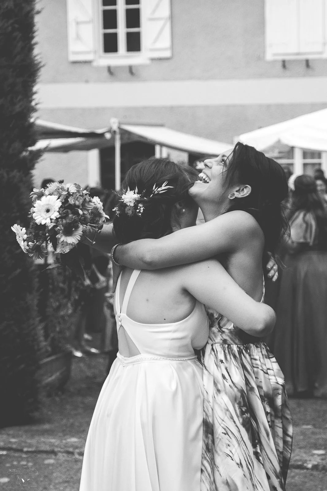 Deux femmes s'embrassent et se sourient dans une ambiance de mariage, une d'elles porte une robe de mariée et un bouquet de fleurs.