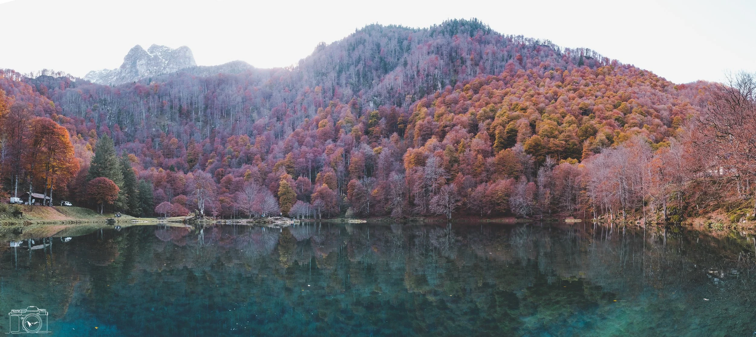 Un lac entouré de forêts aux arbres aux couleurs d'automne, avec des montagnes en arrière-plan, en milieu de journée.