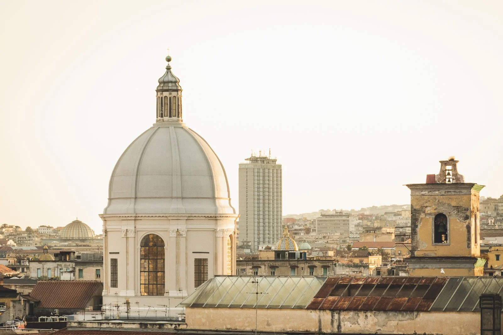 Vue urbaine montrant la coupole d'une église, plusieurs bâtiments anciens et modernes au fond, en fin d'après-midi ou au coucher du soleil.