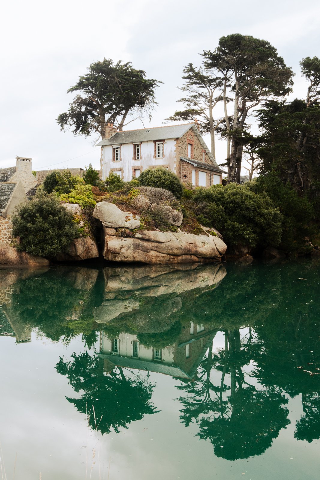 Une maison ancienne en pierre, entourée d'arbres, est reflétée dans l'eau calme d'un lac ou d'une rivière.