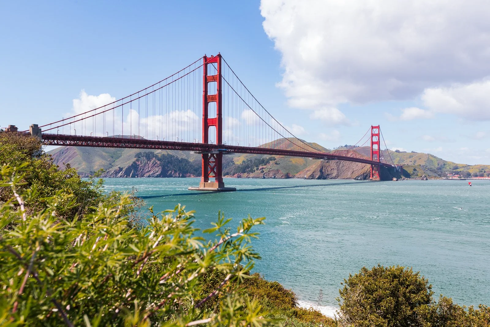 Le pont Golden Gate rouge s'étend au-dessus de la baie de San Francisco, avec des collines en arrière-plan et des buissons verts au premier plan, sous un ciel en partie nuageux.