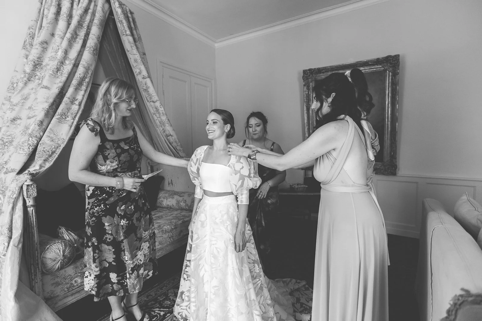 Fête de mariage avec quatre femmes dans une pièce élégante. La mariée sourit, entourée de trois autres femmes, dont une ajuste sa robe, dans une ambiance de préparation nuptiale.
