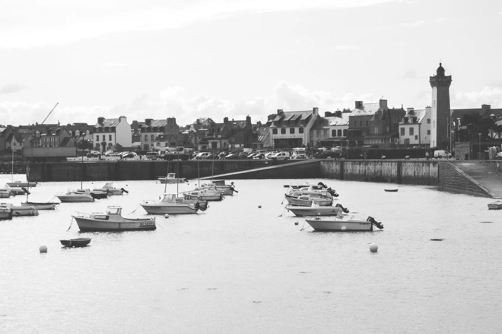 Vue d'un port avec plusieurs bateaux amarrés dans l'eau, en arrière-plan des maisons et un phare. En noir et blanc.