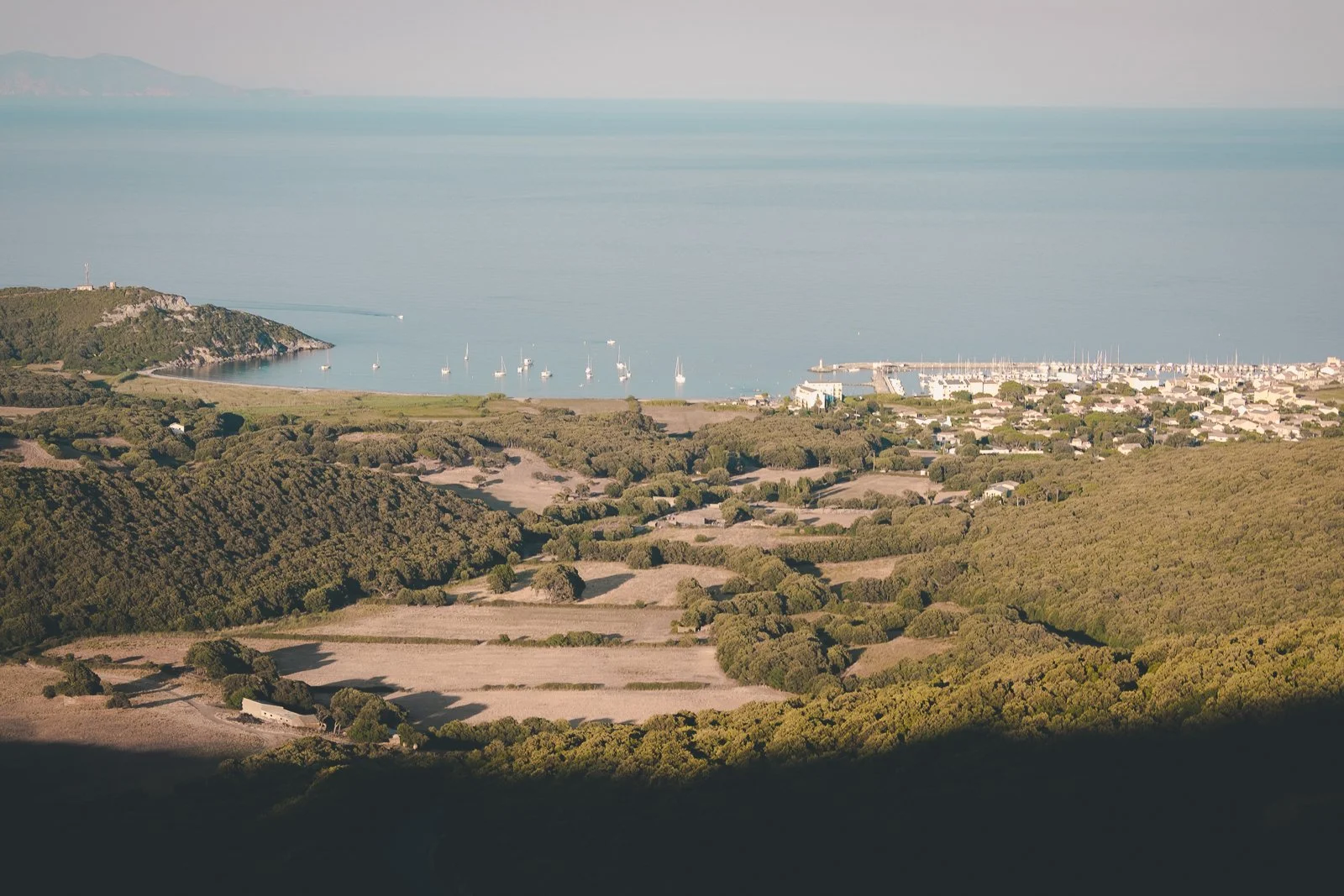 Paysage côtier avec une baie, des bateaux dans l'eau, un port, des maisons, des collines verdoyantes et des montagnes en arrière-plan.