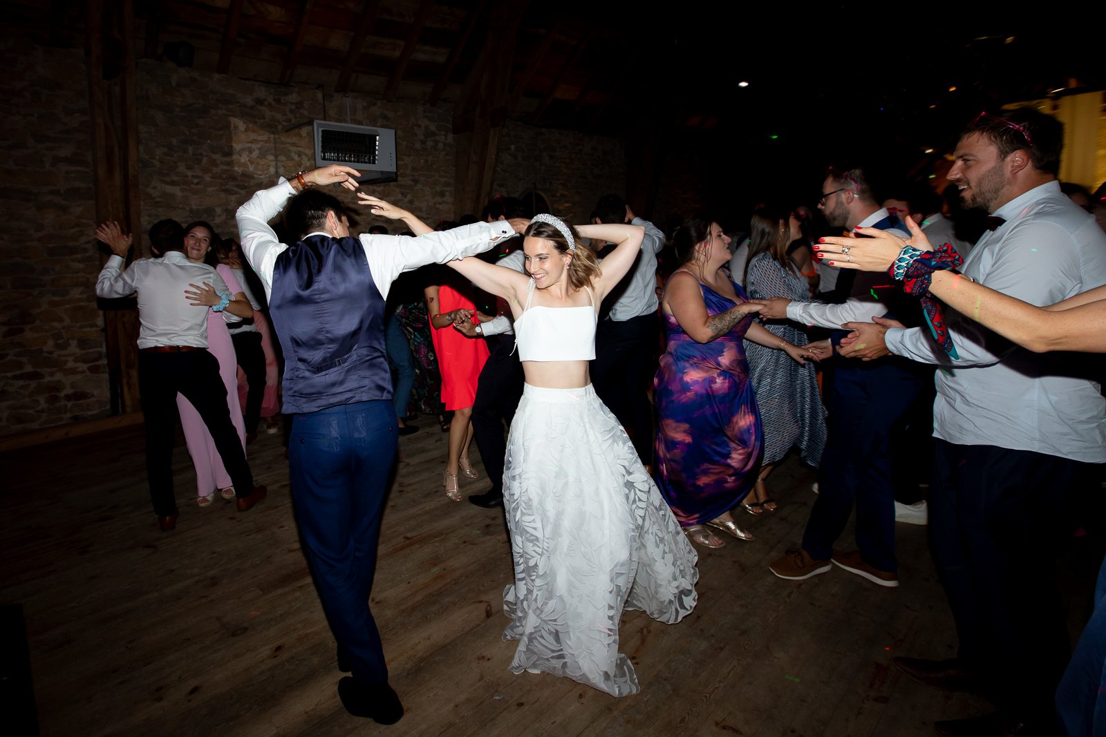 Personne en robe de mariée dansant et souriant entourée de plusieurs autres personnes qui dansent ou applaudissent dans un endroit intimiste avec murs en pierre.